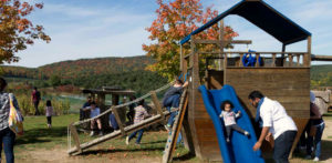 A small child comes down a blue slide attached to a wooden jungle gym shaped like a ship.