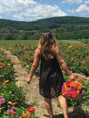 A woman is shown from the back holding a basket of flowers while walking on a path in between rows of zinnias.