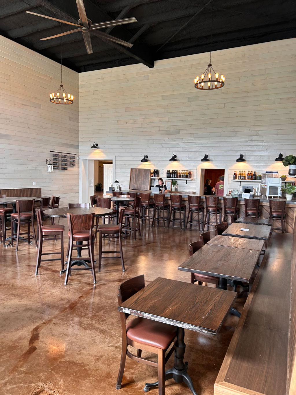 An large open room with deep wood tables with leather seats. Concrete floors are custom stained with warm, rich brown hues with rustic chandelier lighting and white shiplap walls.