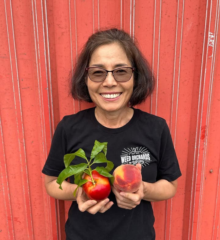 I smiling woman holds up two peaches against a red barn backdrop