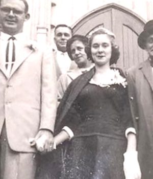 A bride and groom stand outside the church doors holding hands. The bride wearing a black dress and white gloves.
