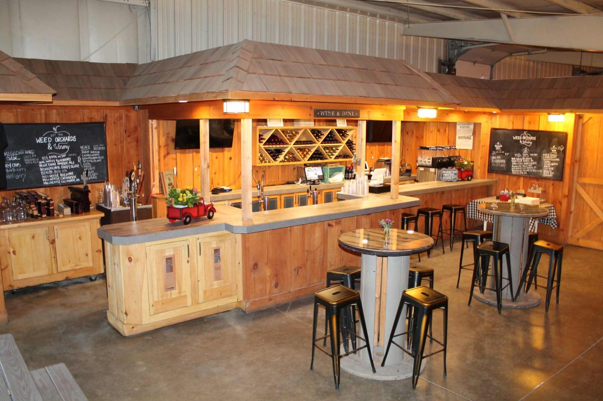 A view from the winery's entrance shows a wooden bar, warm lighting, and tall bistro style tables in the center area